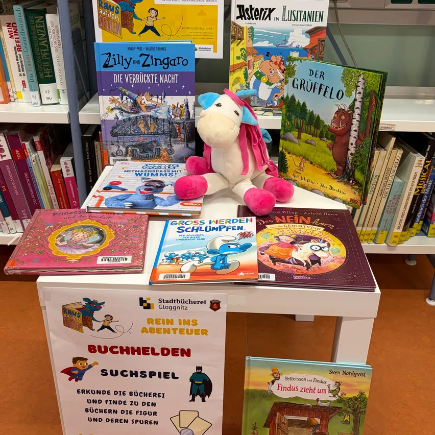 Ein Tisch mit verschiedenen Kinderbüchern, darunter 'Zilly und Zingaro', 'Der Gruffelo' und 'Grossen Schlumpffen', mit einem Plüsch-Einhorn-Spielzeug obenauf.
