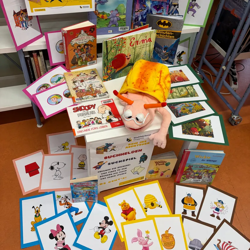 Eine Sammlung von Kinderbüchern und Spielzeug, darunter ein Plüschtier mit gelbem Hut, ist auf einem Holzboden ausgestellt. Die Bücher sind in verschiedenen Farben und Größen und einige haben Comicfiguren darauf. Es gibt auch Karten mit Comicfiguren und eine Box mit der Aufschrift 'Buchhelden Suchspiel'.