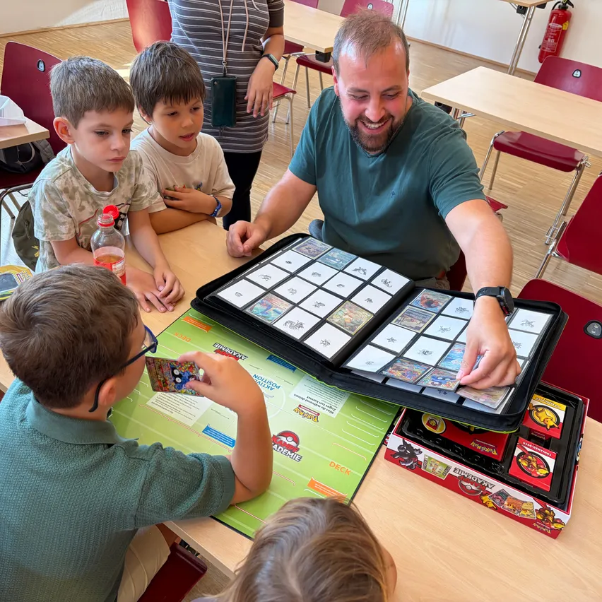 Ein Mann und vier Kinder spielen ein Kartenspiel an einem Tisch. Der Mann lächelt und hält ein Kartenspiel in der Hand.