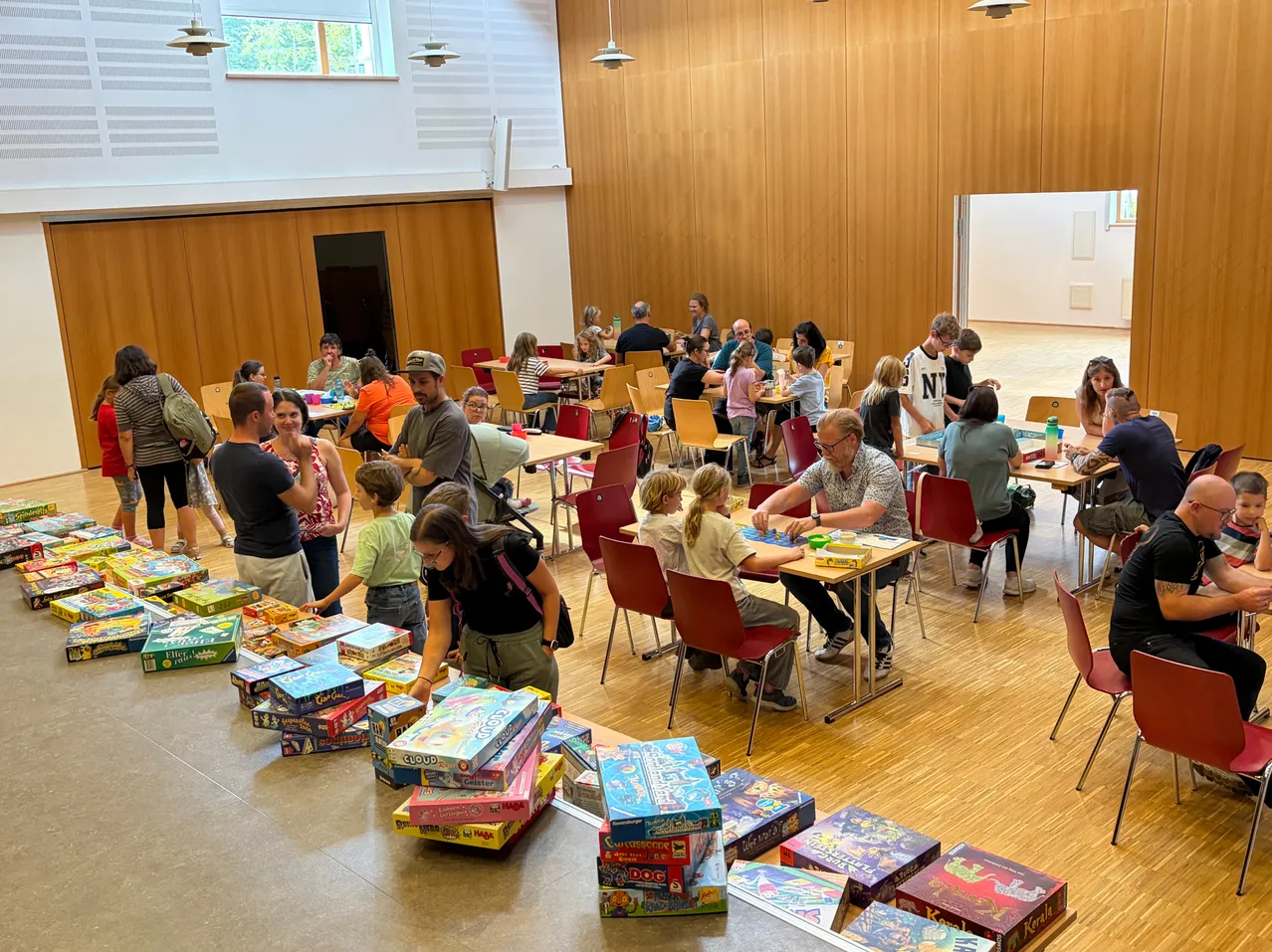 Eine große Gruppe von Menschen versammelt sich in einem Raum, sitzt an Tischen und spielt Brettspiele. Viele Spieleboxen sind auf dem Boden gestapelt.