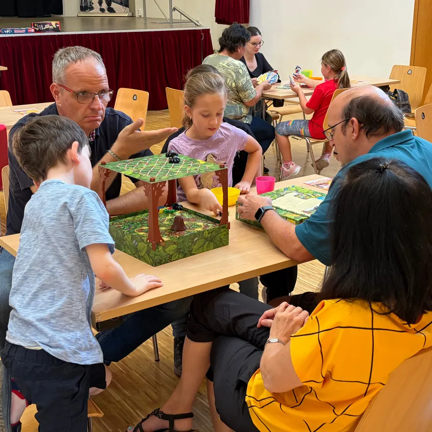 Eine Gruppe von Erwachsenen und Kindern sitzt um einen Tisch herum und spielt ein Brettspiel. Die Kinder sind besonders engagiert, während die Erwachsenen beobachten und gelegentlich interagieren.