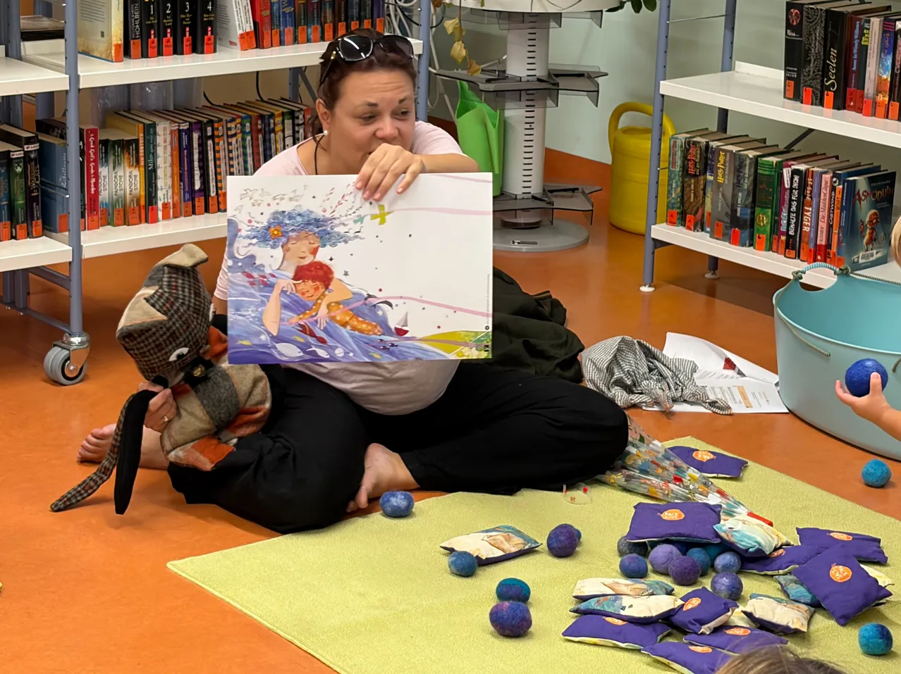 Eine Frau sitzt auf dem Boden und hält ein Buch mit einer farbenfrohen Illustration, umgeben von verschiedenen Bastelmaterialien.