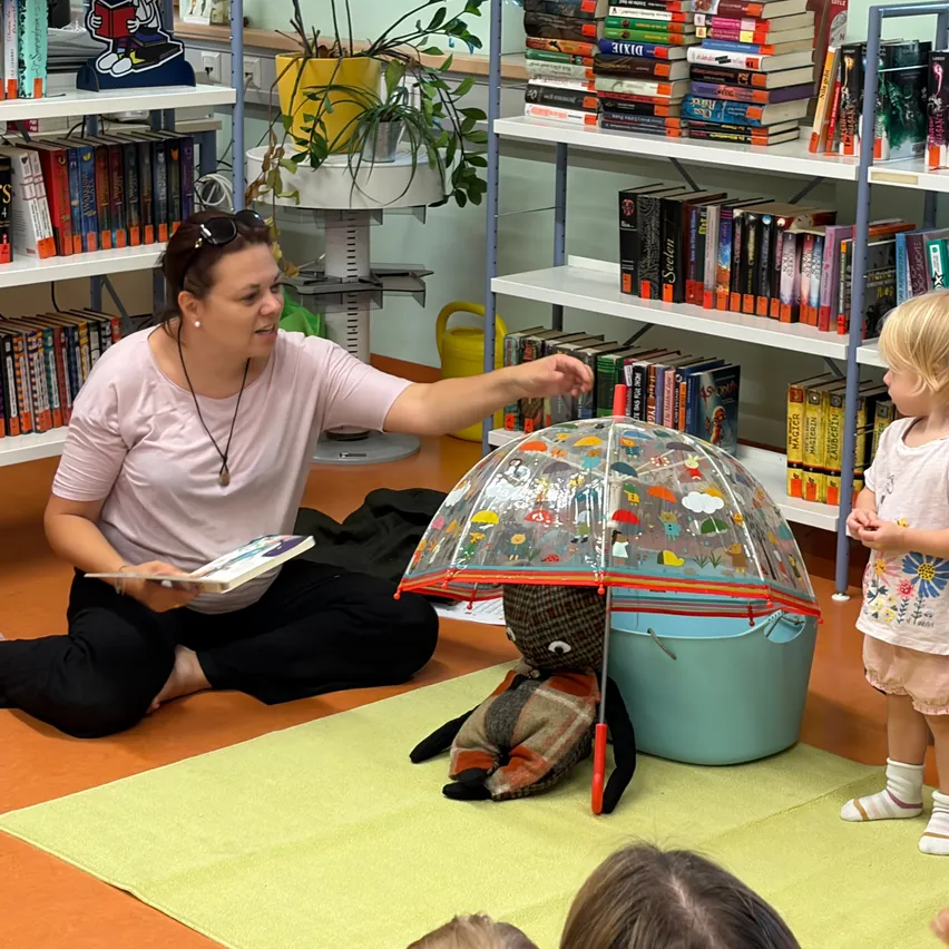 Eine Frau liest einem Kind ein Buch vor, während sie auf dem Boden sitzt, mit einem bunten Regenschirm in der Nähe. Eine Puppe ist unter dem Schirm und Bücherregale im Hintergrund.