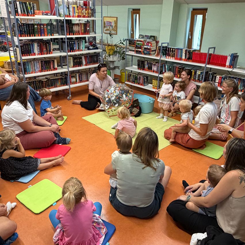 Buchkindergarten: Frauen und Kinder sitzen auf bunten Matten vor Bücherregalen in einer Bibliothek.