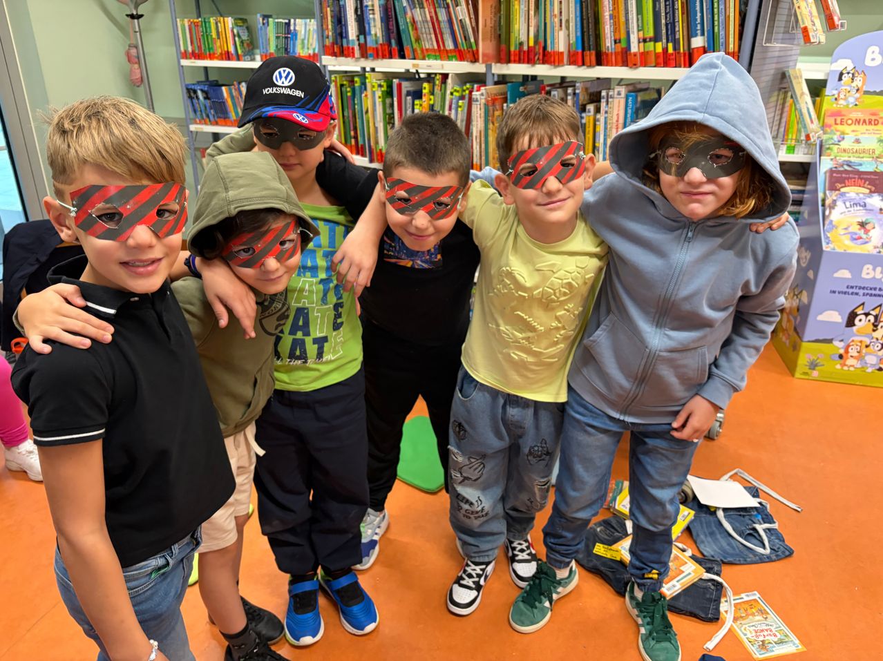 Fünf Jungen mit Masken stehen zusammen in einer Bibliothek. Sie tragen alle verschiedenfarbige Masken. Hinter ihnen befinden sich Regale voller Bücher.