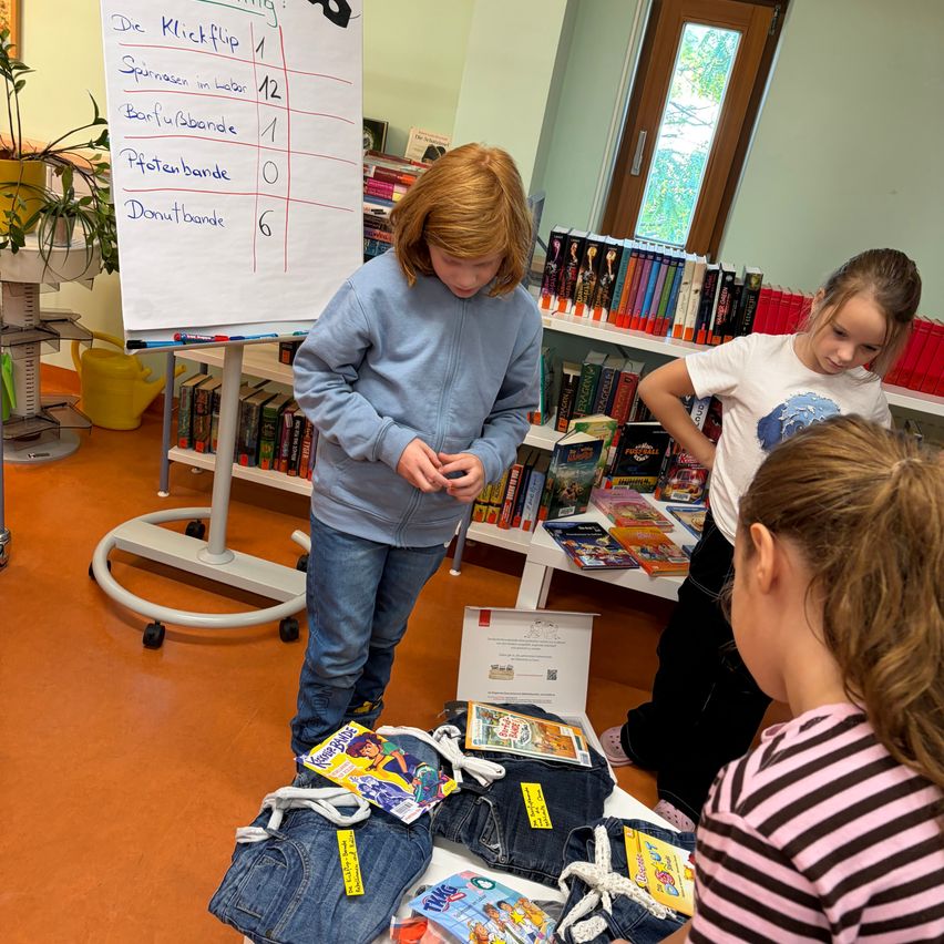 Ein Mädchen in einer Bibliothek, das Bücher in Taschen sortiert, während andere Kinder zusehen. Im Hintergrund steht ein Whiteboard mit einer Liste von Artikeln.