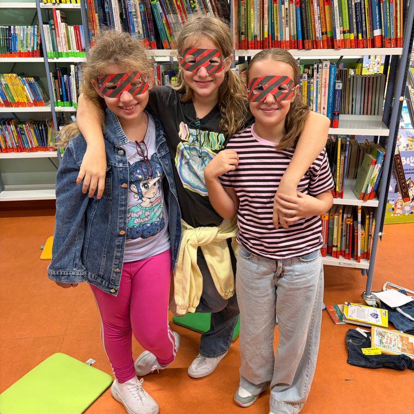 Drei junge Mädchen mit verbundenen Augen und lächelnd, stehen zusammen in einer Bibliothek.