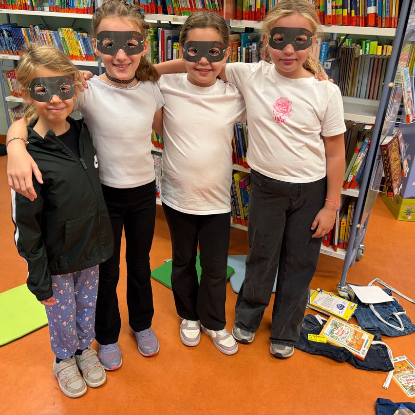 Vier Mädchen mit Masken stehen in einer Bibliothek, lächeln und posieren für ein Foto. Sie sind von Büchern und einer Yogamatte umgeben.