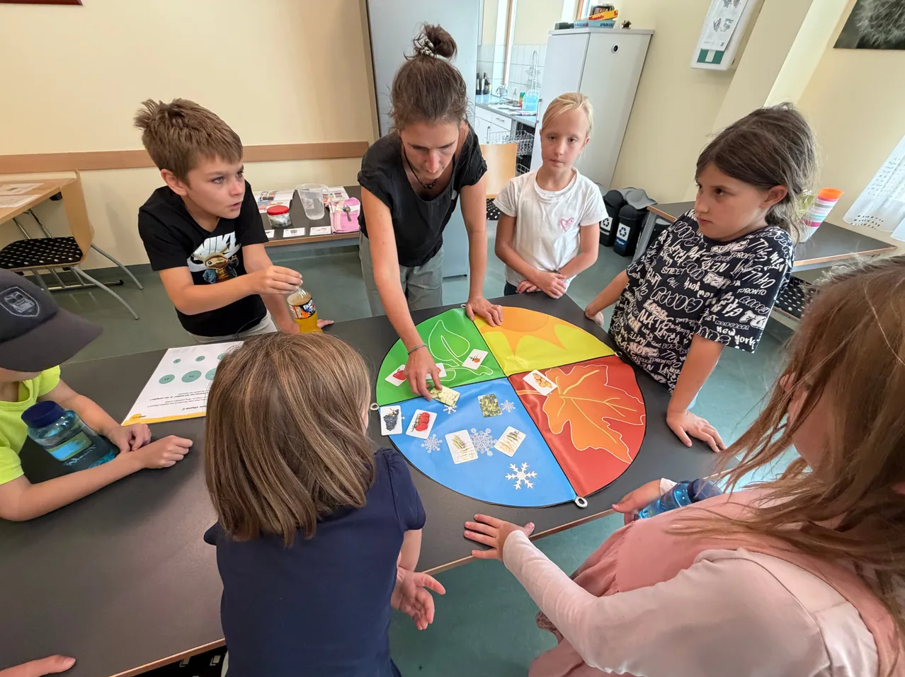 Eine Gruppe von Kindern und einer Frau versammelt sich um einen Tisch in einem Klassenzimmer und beschäftigt sich mit einer Aktivität an einem runden Brett mit verschiedenen farbigen Abschnitten und Karten.