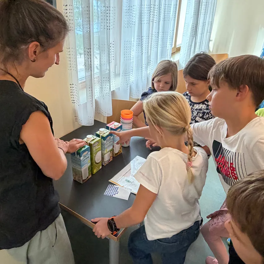 Eine Frau mit einer Halskette steht hinter einem Tisch mit Kindern. Die Kinder schauen auf den Tisch und scheinen eine Aktivität zu machen.