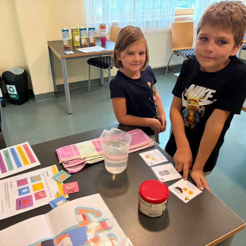 Zwei Kinder stehen neben einem Tisch mit verschiedenen Gegenständen darauf. Ein Mädchen trägt ein blaues Shirt mit Blumendesign und ein Junge trägt ein schwarzes Shirt mit einem Cartoon-Charakter darauf.
