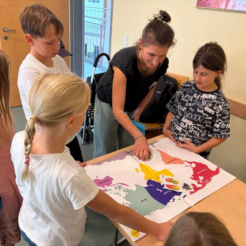 Eine Gruppe von Kindern und einer erwachsenen Frau versammelt sich um einen Tisch und betrachtet eine große Weltkarte. Die Kinder zeigen auf verschiedene Länder und diskutieren etwas.
