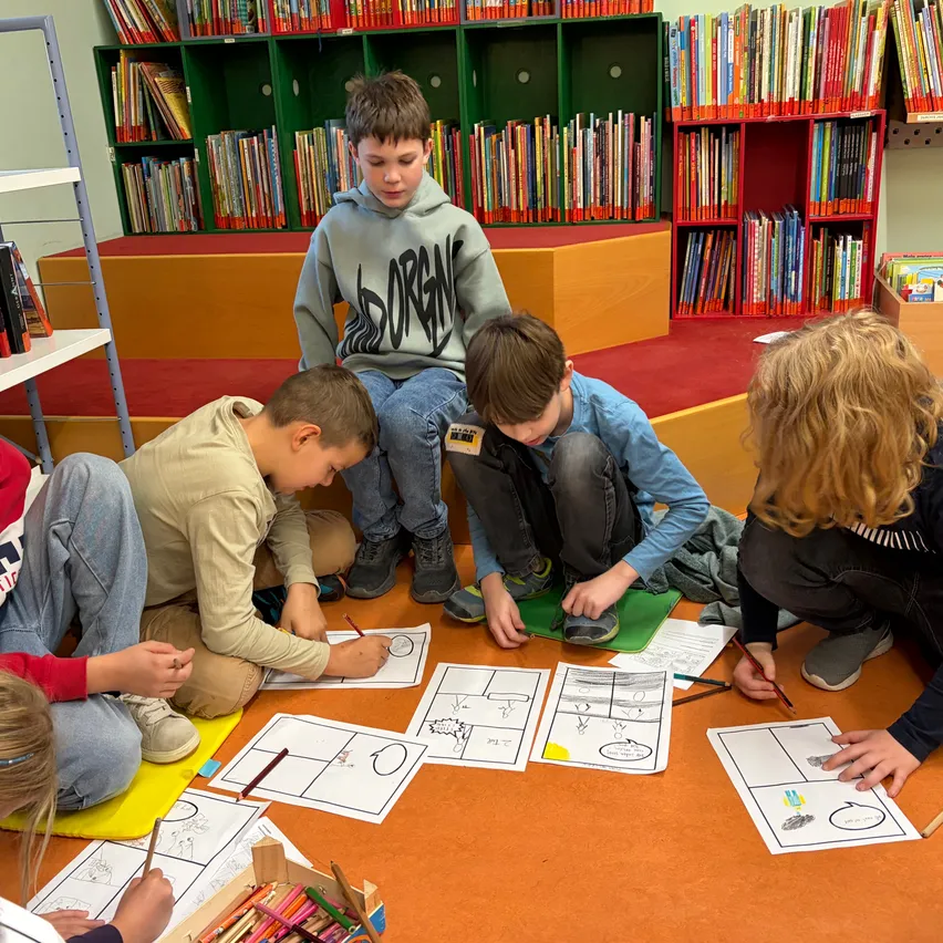 Eine Gruppe von Kindern sitzt auf dem Boden in einer Bibliothek und zeichnet mit Bleistiften auf Papier.