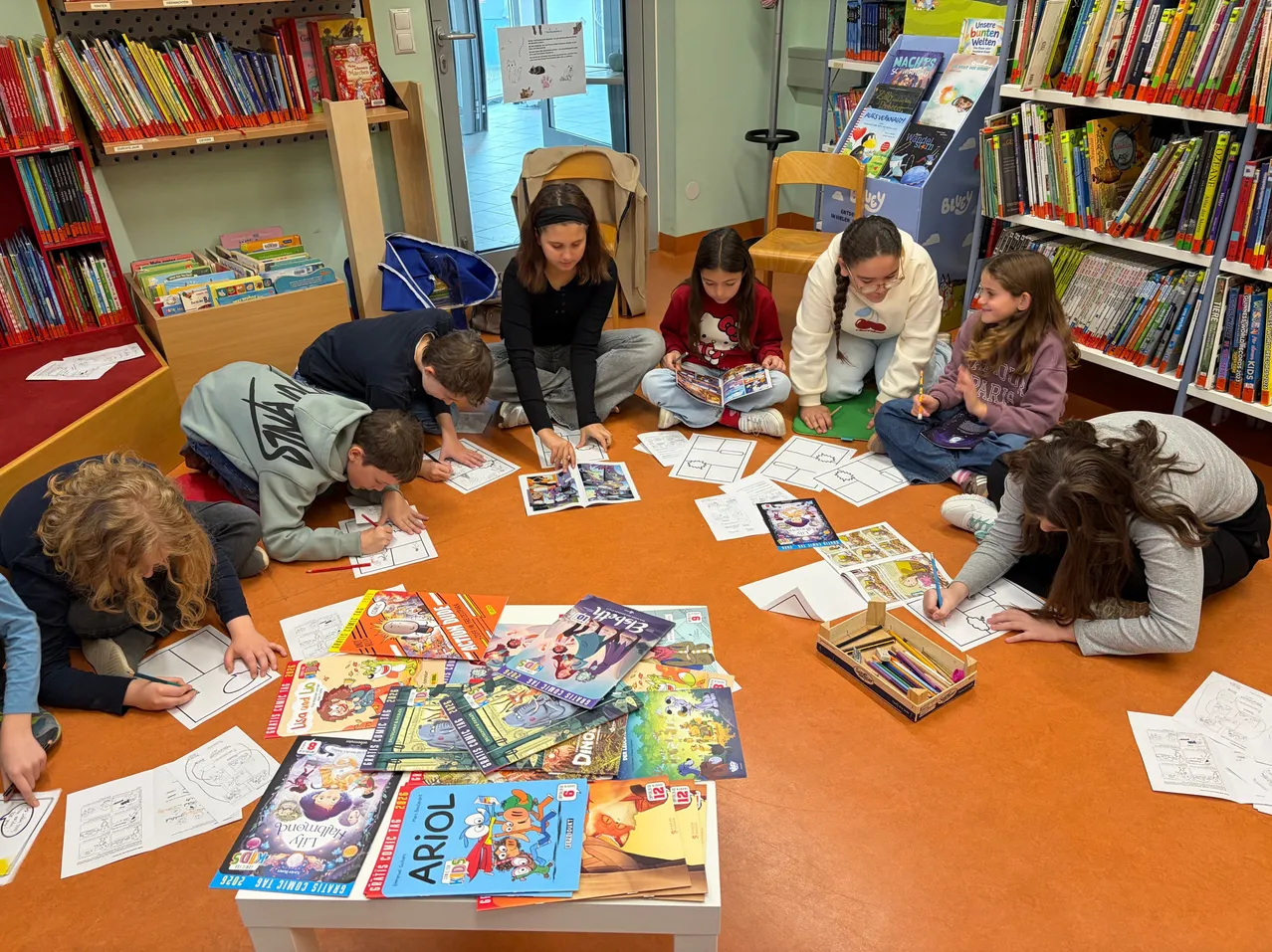 Eine Gruppe von Kindern sitzt auf dem Boden in einer Bibliothek, zeichnet auf Papier und in Malbüchern. Eine Frau sitzt unter ihnen, und sie sind von Büchern und Papieren umgeben.