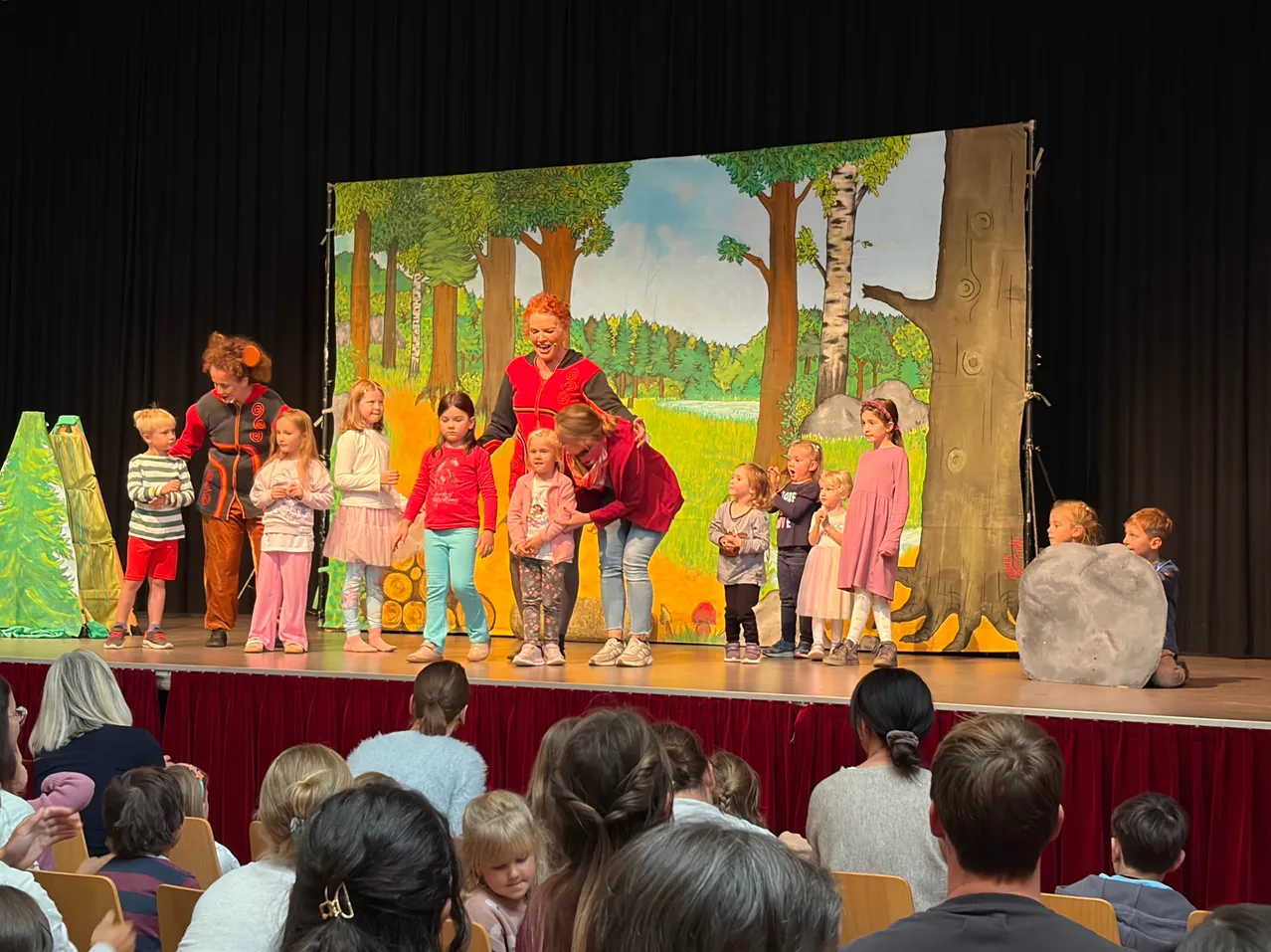 Eine Gruppe von Kindern und Erwachsenen steht auf der Bühne, mit einem gemalten Hintergrund eines Waldes. Die Kinder sind in verschiedenen Kostümen gekleidet und stehen in einer Reihe.