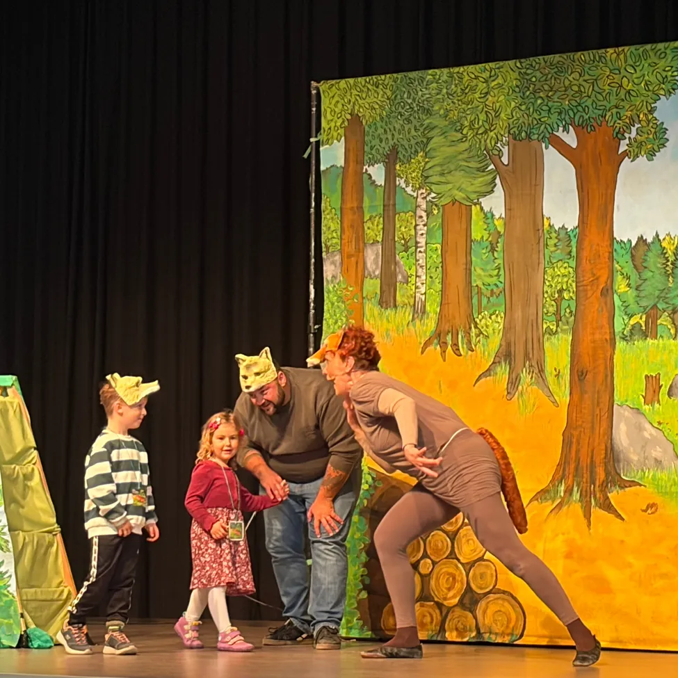 Eine Gruppe von Menschen steht auf der Bühne und führt ein Theaterstück auf. Ein Mann und eine Frau stehen in der Mitte, und zwei Kinder stehen auf beiden Seiten von ihnen. Die Bühne ist mit einem Hintergrund eines Waldes dekoriert.