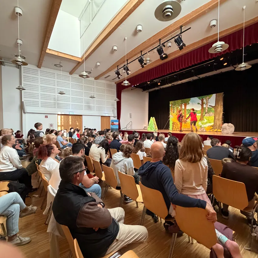 Eine große Gruppe von Menschen sitzt in einem Theater auf Stühlen und schaut sich eine Aufführung auf der Bühne an.