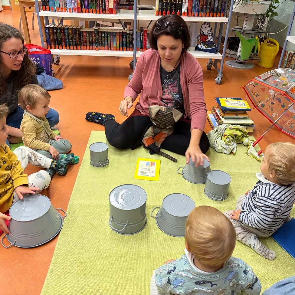 Eine Gruppe junger Kinder und zwei Erwachsene sitzen auf dem Boden in einer Bibliothek und beschäftigen sich mit einer musikalischen Aktivität mit Metalleimern.