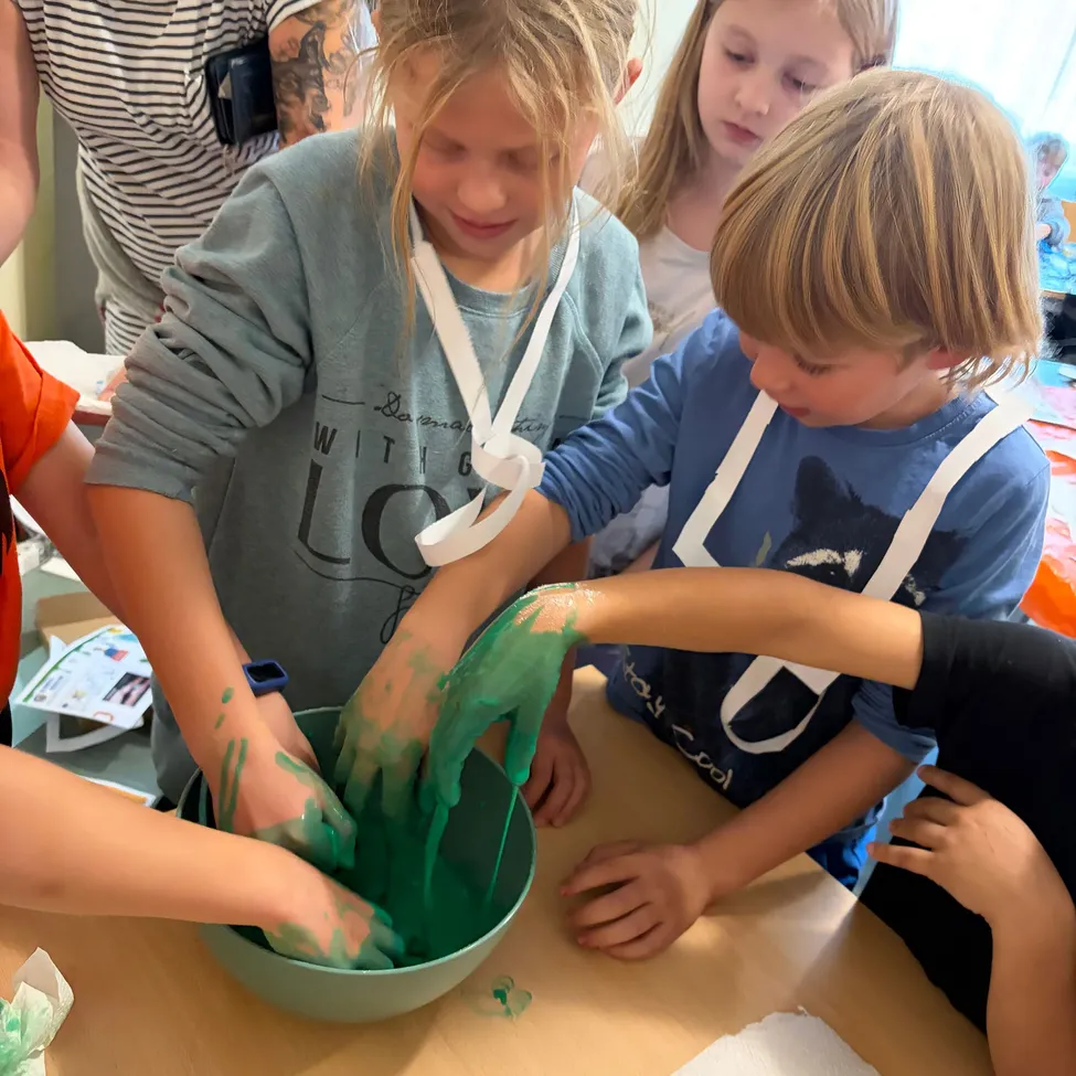 Eine Gruppe von Kindern versammelt sich um einen Tisch, jeder mit den Händen in einer Schüssel mit grünem Schleim.