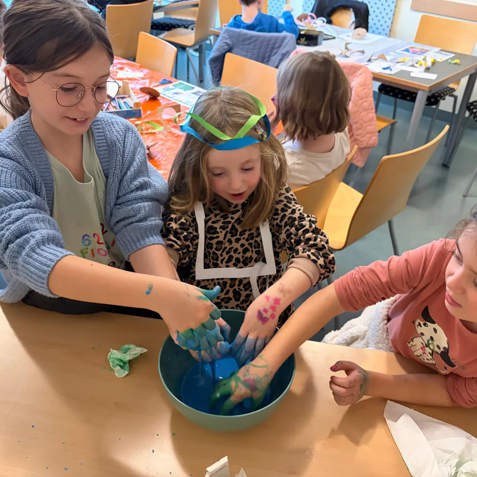 Drei Mädchen sitzen an einem Tisch in einem Klassenzimmer. Das Mädchen auf der linken Seite trägt eine Brille und hat blaue Farbe auf den Händen. Das Mädchen in der Mitte trägt eine Stirnband und hat rosa Farbe auf den Händen. Das Mädchen auf der rechten Seite trägt ein rosa T-Shirt und hat blaue Farbe auf den Händen.