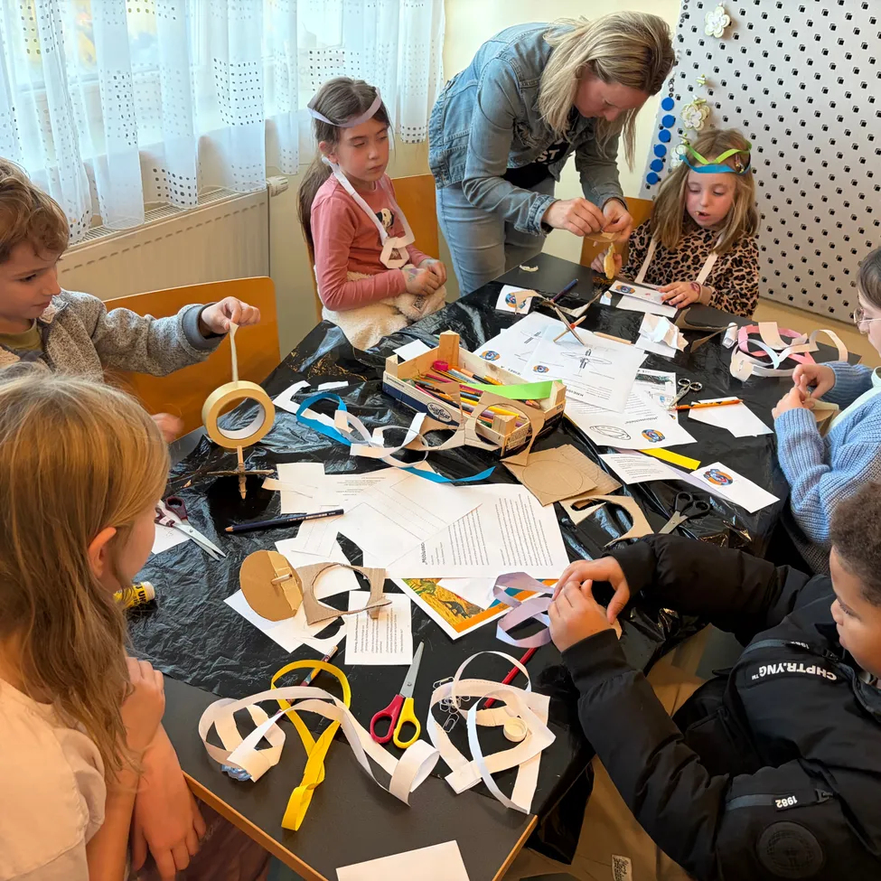 Eine Gruppe von Kindern und einem Erwachsenen sitzen um einen Tisch herum und beschäftigen sich mit einer Bastelaktivität. Der Tisch ist mit einem schwarzen Tuch bedeckt und hat verschiedene Bastelmaterialien wie Scheren, Klebeband und Papier. Die Kinder tragen Schürzen und Stirnbänder, und der Erwachsene hilft ihnen.
