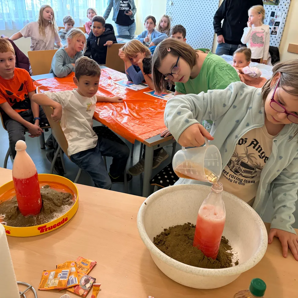 Eine Gruppe von Kindern sitzt um einen Tisch in einem Klassenzimmer. Ein Mädchen gießt eine Flüssigkeit aus einer Flasche in eine Schüssel mit Sand.