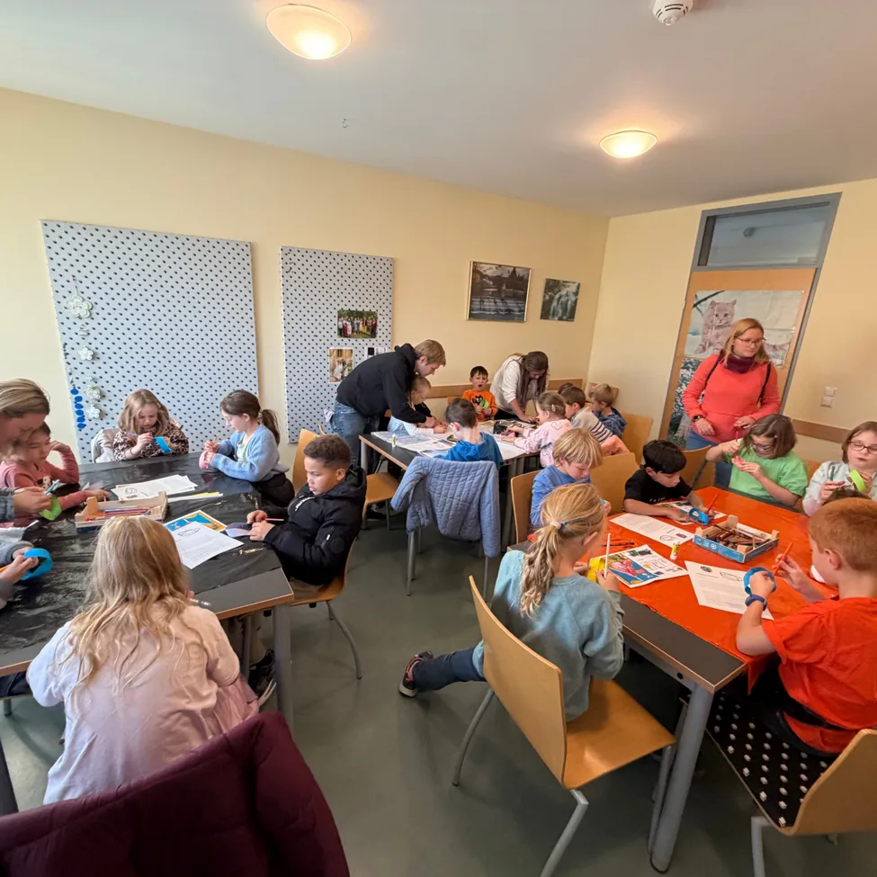 Eine Gruppe von Kindern sitzt an Tischen in einem Klassenzimmer und ist mit einer Aktivität beschäftigt. Zwei Erwachsene sind anwesend, einer steht und der andere sitzt.