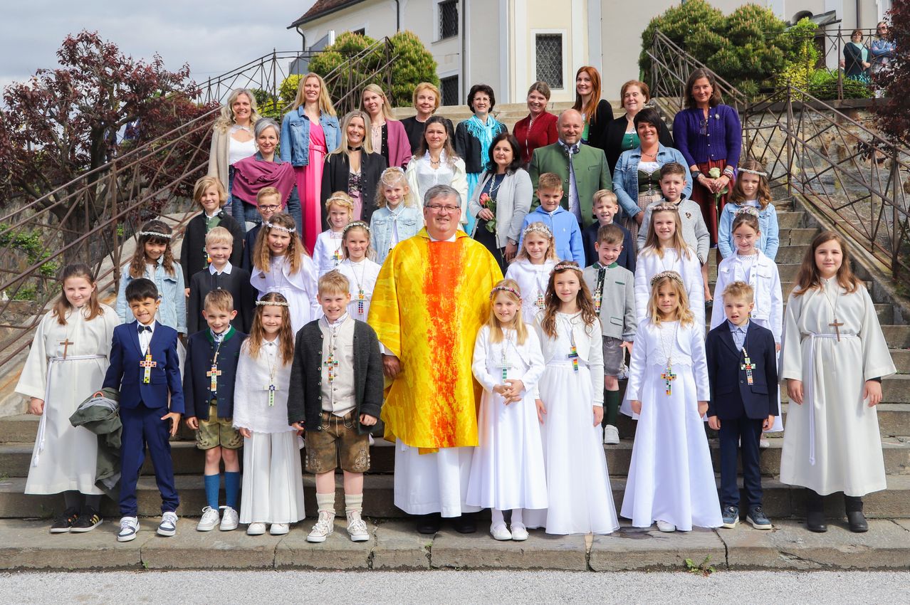 Ein Priester mit Brille posiert mit einer Gruppe von Menschen, darunter Kinder, auf den Stufen einer Kirche.