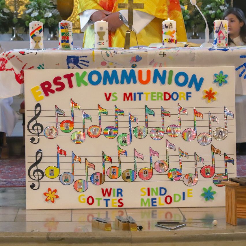 Ein Priester steht an einem Altar mit einem großen Banner, das 'Erstkommunion vs. Mitterdorf' besagt. Das Banner zeigt Musiknoten und Flaggen mit dem Text 'Wir sind Gottes Melodie'.