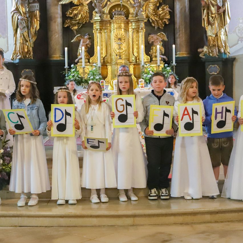 Eine Gruppe von Kindern in weißen Kleidern, die Musiknoten halten, steht vor einem Altar. Die Kinder halten Musiknoten mit den Buchstaben G, D, F, A, C, E und H.