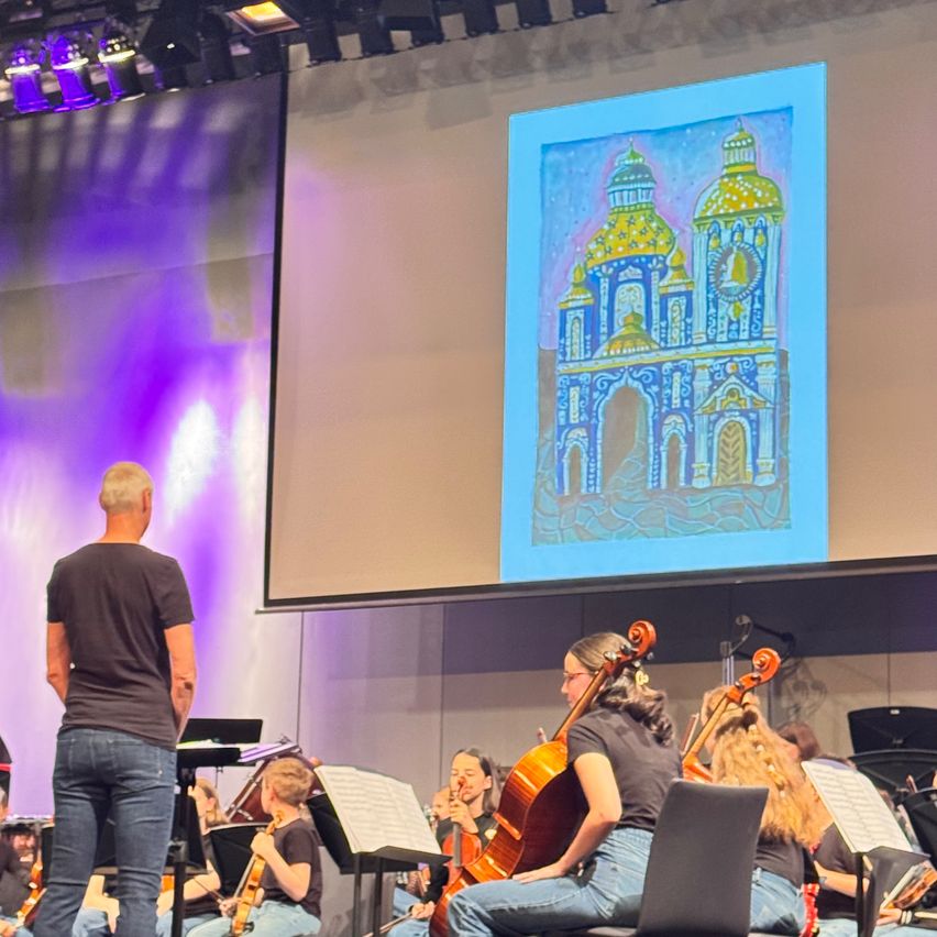 Eine Band spielt auf der Bühne, während eine Leinwand eine farbenfrohe Zeichnung eines Gebäudes zeigt. Der Dirigent steht auf der linken Seite und die Musiker sitzen auf Stühlen.