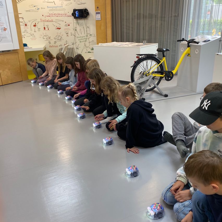 Eine Gruppe von Kindern sitzt auf dem Boden, jedes interagiert mit einem kleinen Roboter, während ein gelbes Fahrrad in der Nähe steht. Der Raum hat eine Tafel mit Zeichnungen und eine an der Wand montierte Projektionsleinwand.