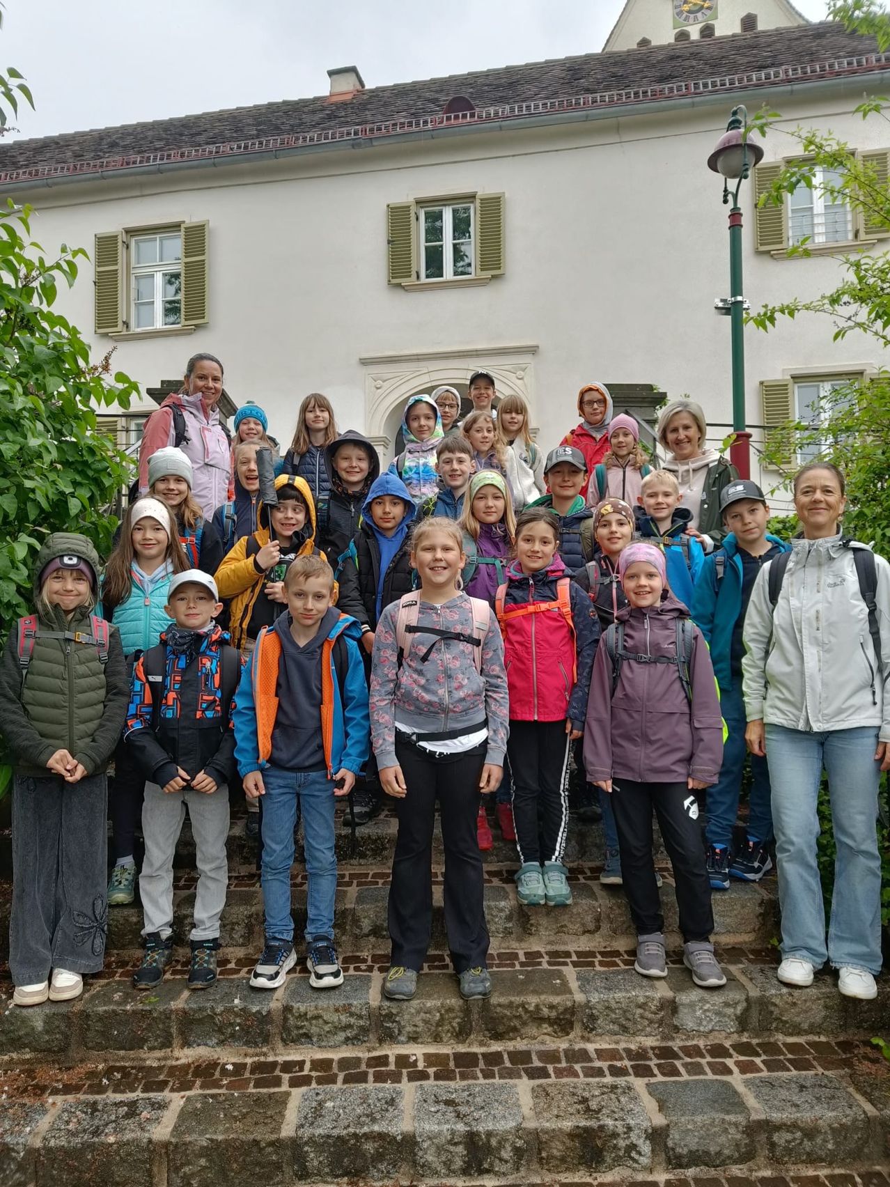 Eine Gruppe von Kindern und Erwachsenen, in Winterkleidung gekleidet, posiert für ein Foto vor einem Gebäude mit mehreren Fenstern und einem grünen Laternenpfahl.