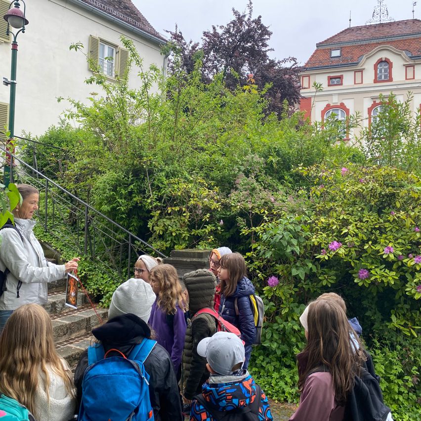 Eine Gruppe von Kindern und einem Erwachsenen versammelt sich auf den Stufen vor einem Gebäude mit einem Garten. Sie hören der Frau zu, die ein Buch hält.