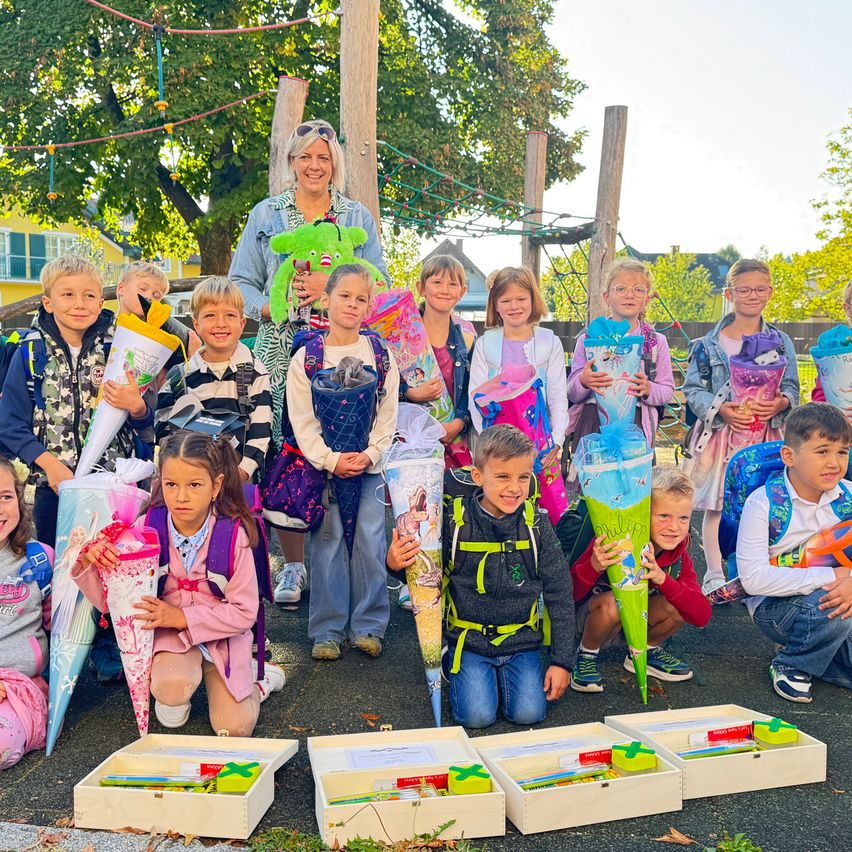 Eine Gruppe von Kindern und eine erwachsene Frau posieren für ein Foto draußen. Sie halten Geschenktüten in den Händen und stehen vor Aufbewahrungsboxen auf dem Boden. Sie alle lächeln und sehen fröhlich aus. Hinter ihnen sind Bäume und ein Spielplatz.