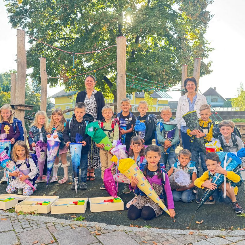 Eine Gruppe von Kindern und zwei Frauen stehen und sitzen auf einem Ziegelboden und halten Geschenke. Sie alle lächeln und posieren für ein Foto. Dahinter stehen Holzpfosten und ein Baum. In der Ferne sind Häuser und Bäume zu sehen.