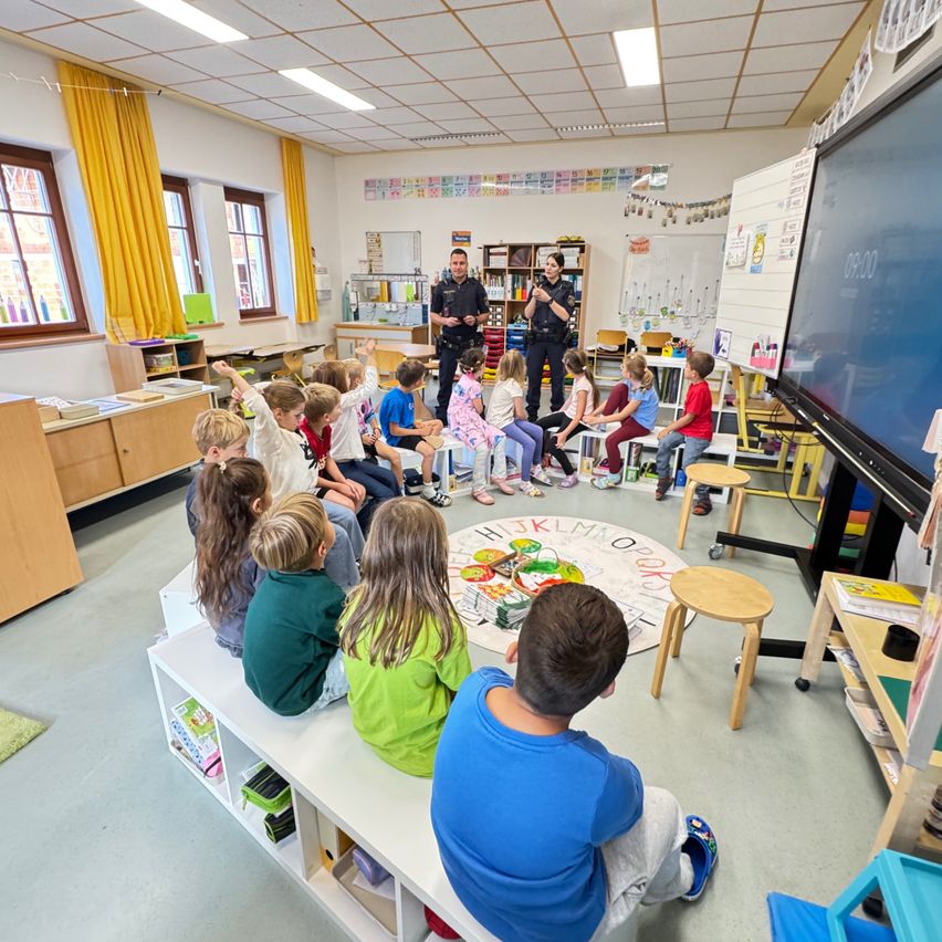 Grundschulkinder sitzen in einem Klassenzimmer in einem Kreis um zwei Polizeibeamte. Die Polizeibeamten sprechen mit den Schülern, während ein großer Monitor an der Wand hängt.