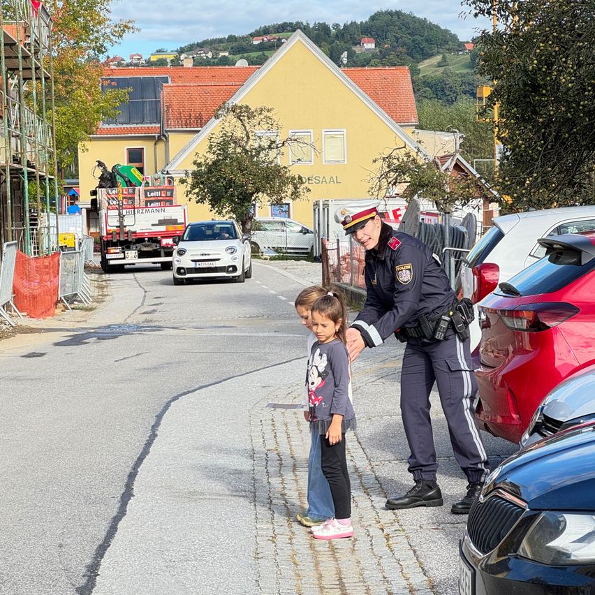 Zwei Kinder stehen auf einer Straße, die von einem Polizisten geführt werden. Dahinter fährt ein Feuerwehrwagen vorbei. Im Bereich sind geparkte Autos, Gebäude und eine Baustelle.