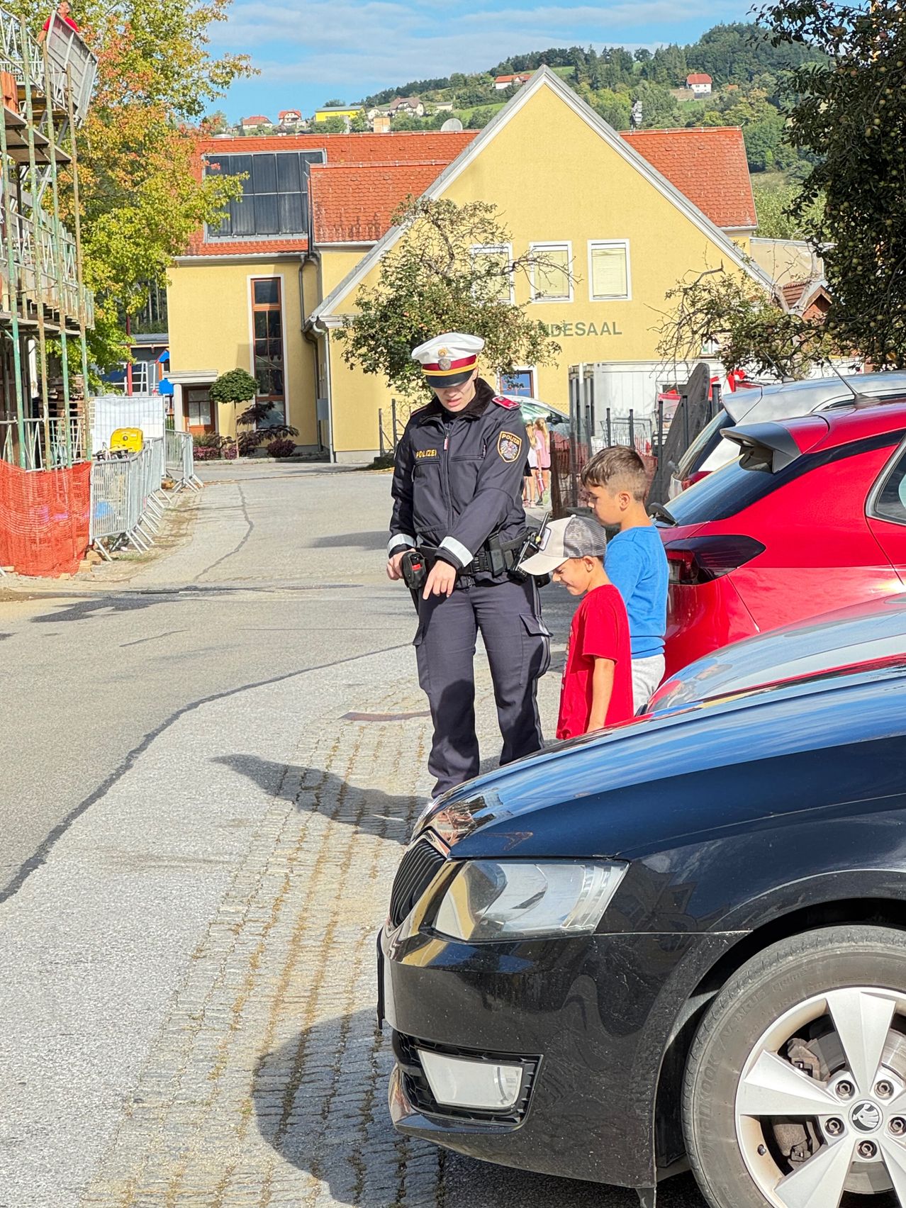 Ein Polizist steht auf der Straße und unterhält sich mit zwei Jungen vor einem gelben Gebäude und geparkten Autos.