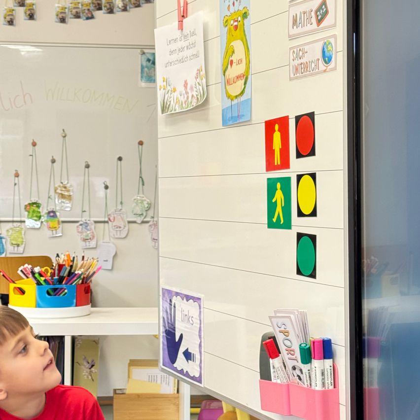 Ein Kind in einem Klassenzimmer schaut an einer Pinnwand mit verschiedenen pädagogischen Postern und farbigen Symbolen nach oben. Im Hintergrund befindet sich ein Schreibtisch mit einem bunten Stifthalter und Spielzeug.