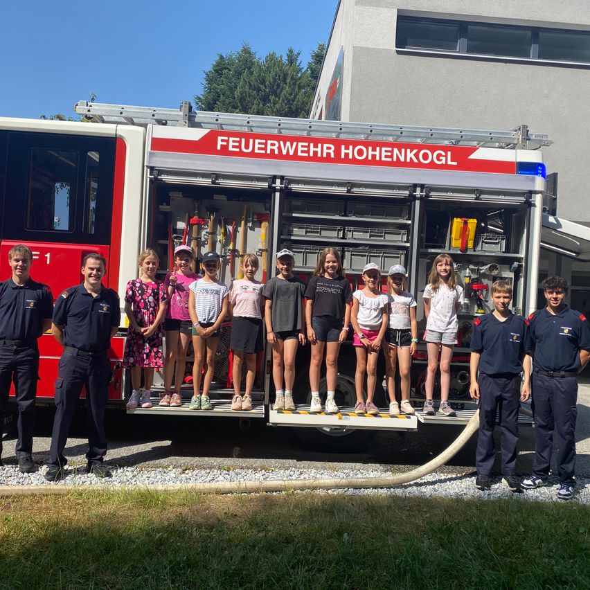 Eine Gruppe von Kindern steht lächelnd auf den Stufen eines Feuerwehrwagens. Zwei Feuerwehrleute sind in der Nähe, einer hält eine Feuerwehrschläuche. Der Wagen ist vor einem Gebäude geparkt.