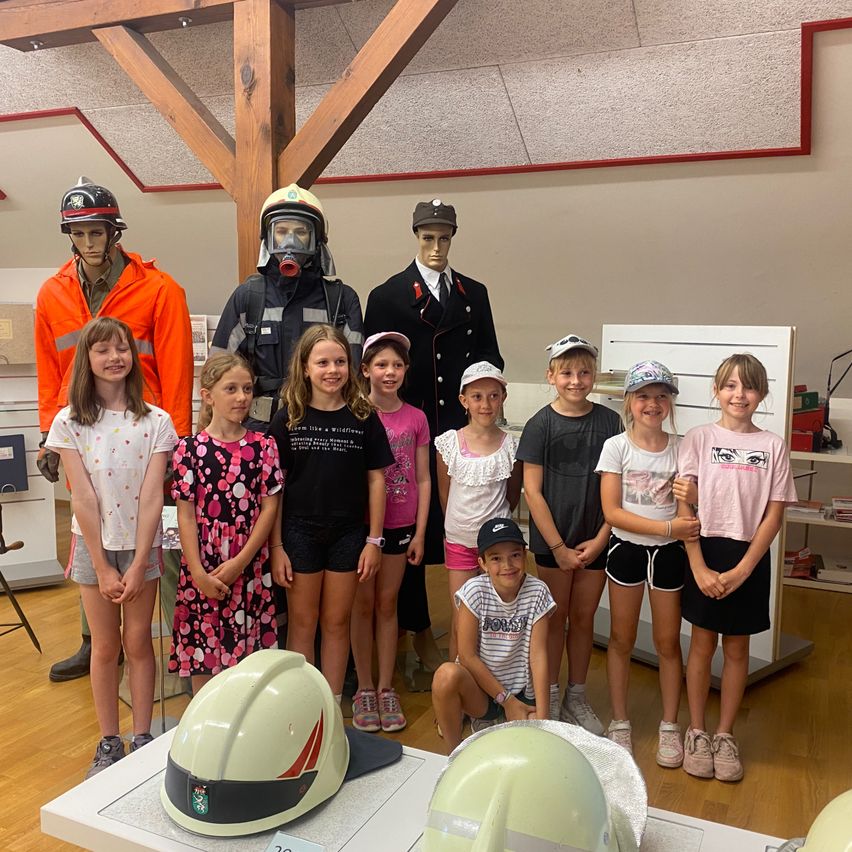 Eine Gruppe junger Mädchen und Jungen steht neben zwei Feuerwehrmannequins und Helmen auf einem Tisch. Sie befinden sich in einem Raum mit Holzböden und weißen Wänden.