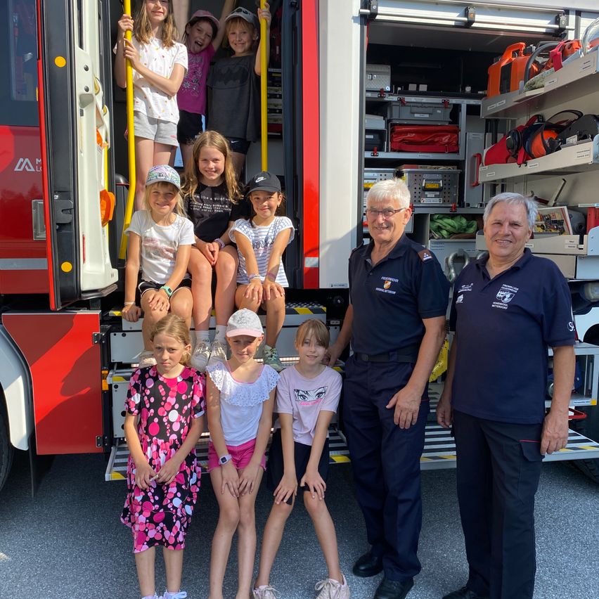 Eine Gruppe lächelnder Kinder und zwei Männer stehen vor einem Feuerwehrauto, einige sitzen auf den Stufen. Die Männer tragen Uniformen und Brillen, und das Fahrzeug ist mit Ausrüstung gefüllt.
