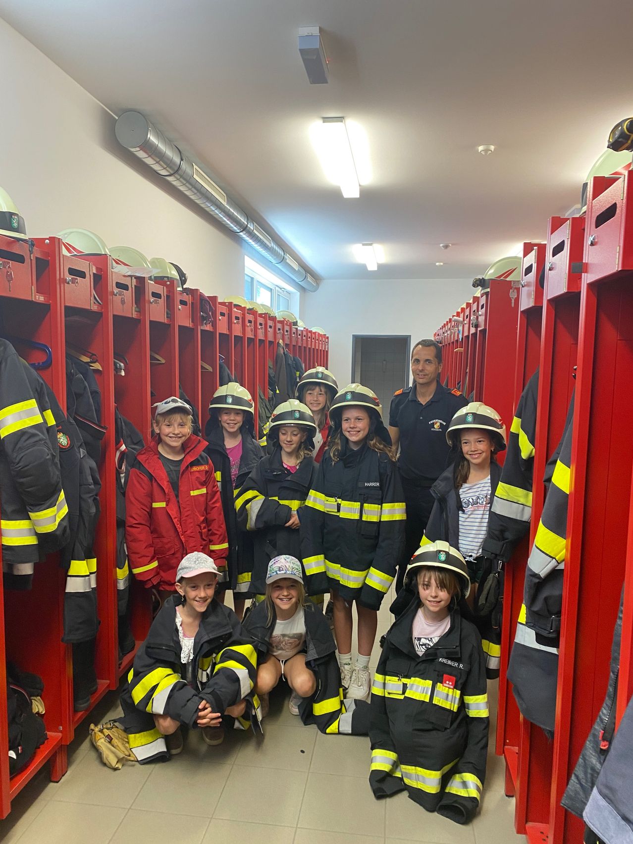 Eine Gruppe von Kindern in Feuerwehruniformen steht in einer Umkleidekabine mit roten Spinden. Ein Mann in Uniform steht hinter ihnen.