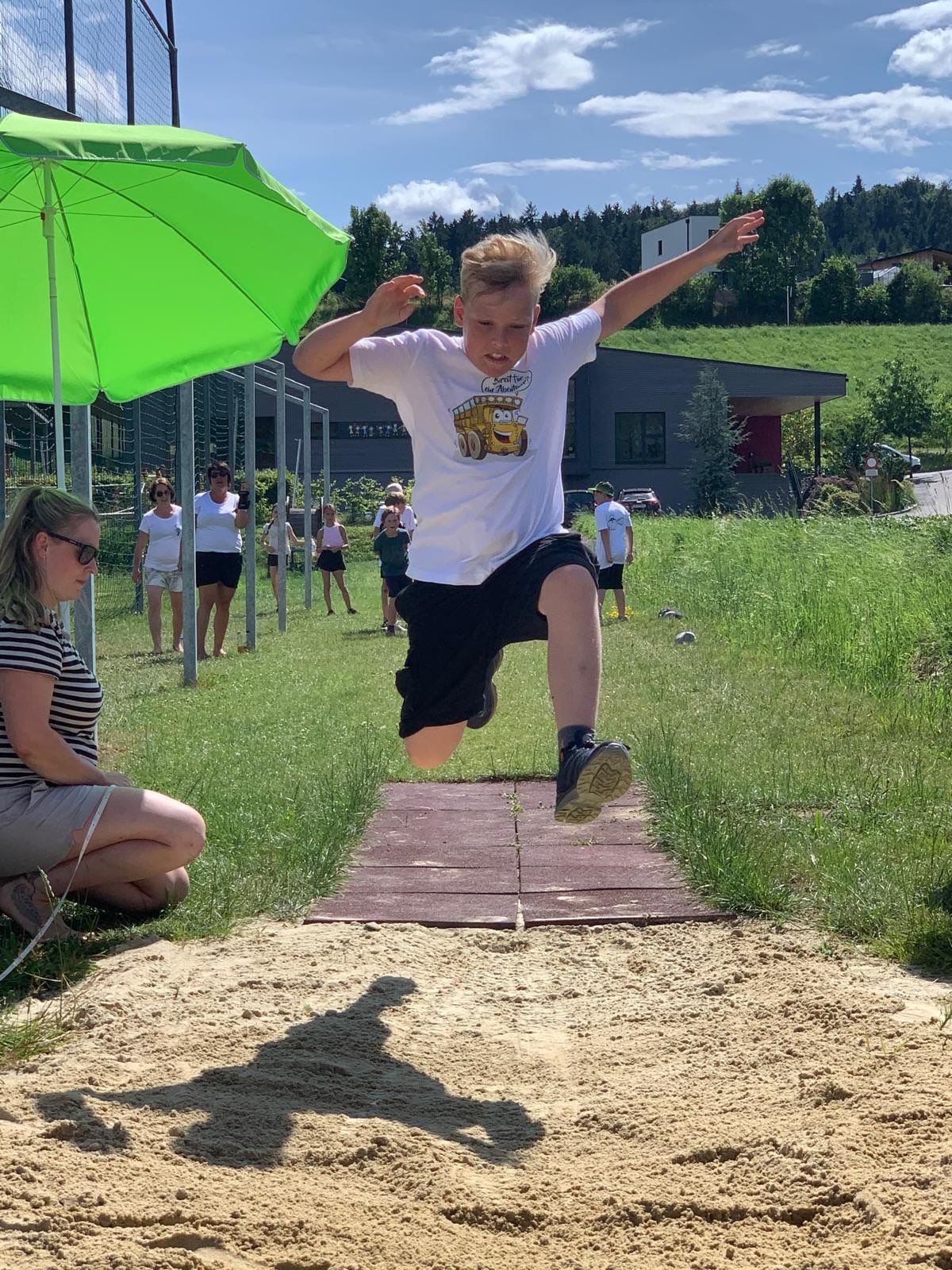 Ein junger Junge springt auf einer Freiluftveranstaltung in eine Sandgrube, mit einem grünen Schirm im Hintergrund. Mehrere Menschen stehen hinter ihm und beobachten.