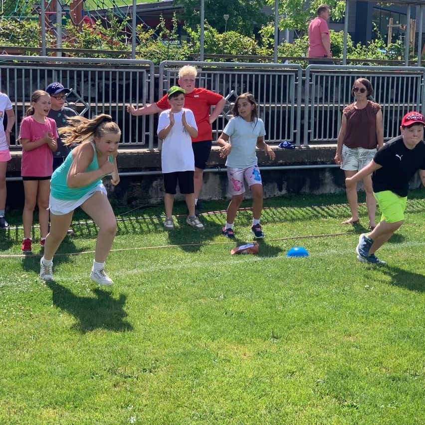 Eine Gruppe von Kindern spielt ein Spiel mit einem blauen Frisbee auf einem Rasenfeld. Eine Frau und ein Mann stehen hinter einem Metallzaun und beobachten.