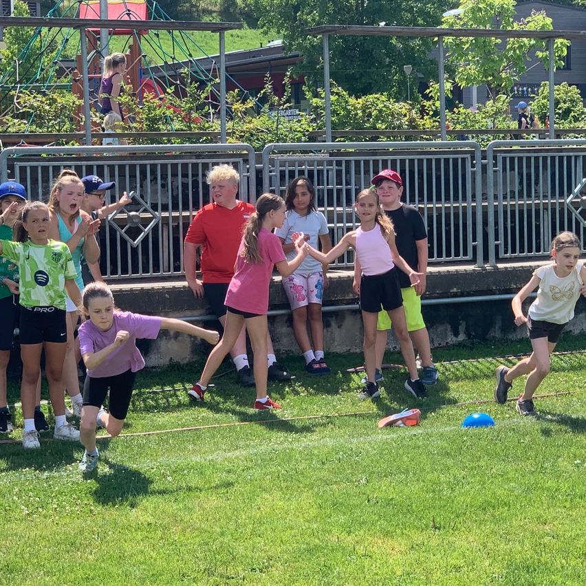 Eine Gruppe von Kindern spielt draußen. Sie laufen und halten sich an den Händen, während einige hinter einem Zaun zuschauen. Im Hintergrund ist ein Spielplatz zu sehen.