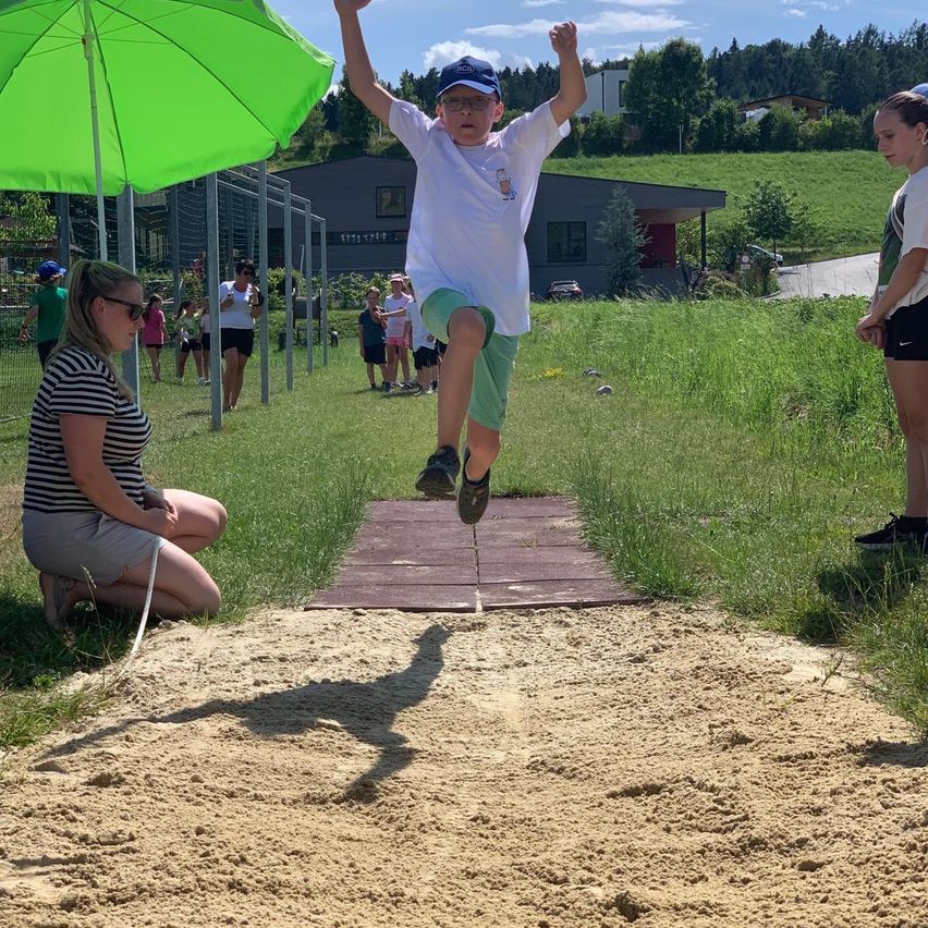Ein Junge springt in eine Sandgrube. Eine erwachsene Frau beobachtet. Ein Mädchen steht vor einem grünen Schirm. Dahinter steht eine Gruppe von Menschen und beobachtet. Im Hintergrund sind Gebäude und eine Straße zu sehen.