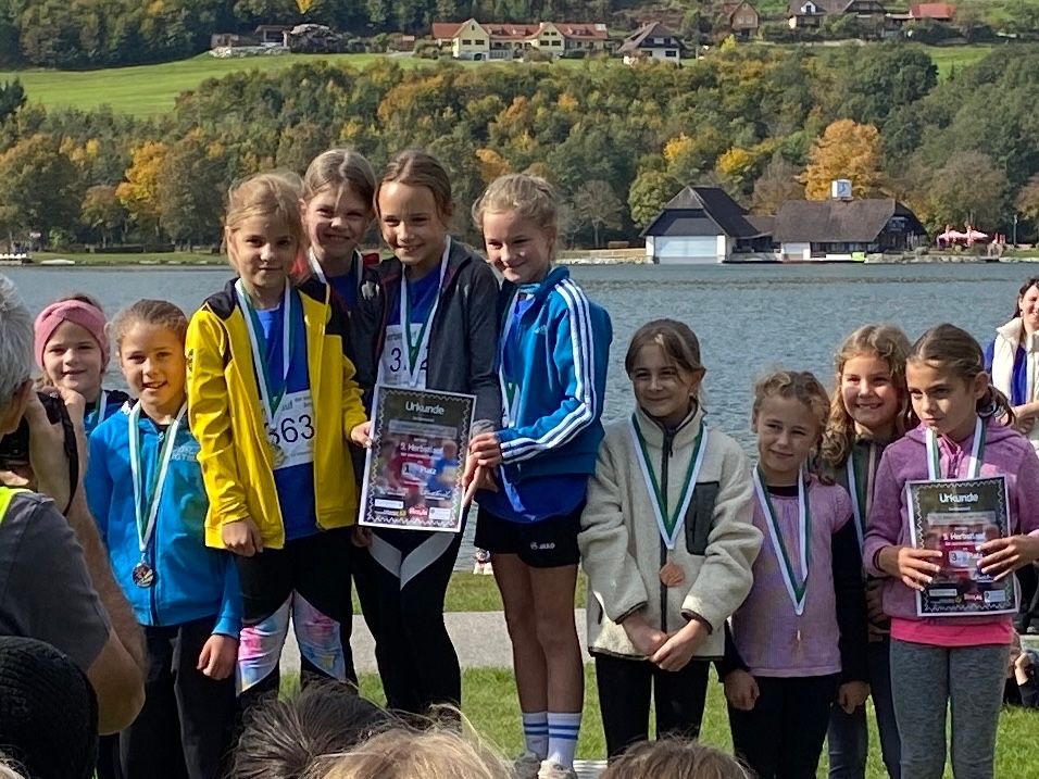Eine Gruppe junger Mädchen steht zusammen am See, trägt Medaillen, lächelt und hält eine Urkunde. Bäume und Häuser sind im Hintergrund zu sehen.