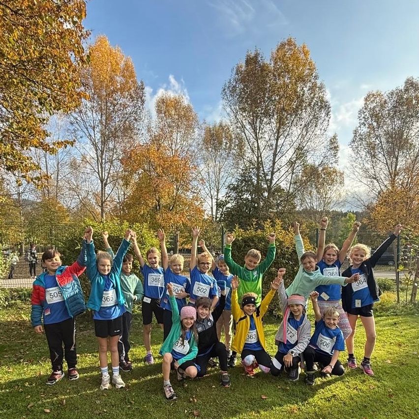 Eine Gruppe von Kindern in bunten Laufoutfits posiert für ein Foto auf einem Rasenfeld mit Bäumen im Herbst.